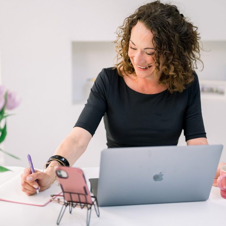 A woman with curly hair works on a laptop, smiling while taking notes.